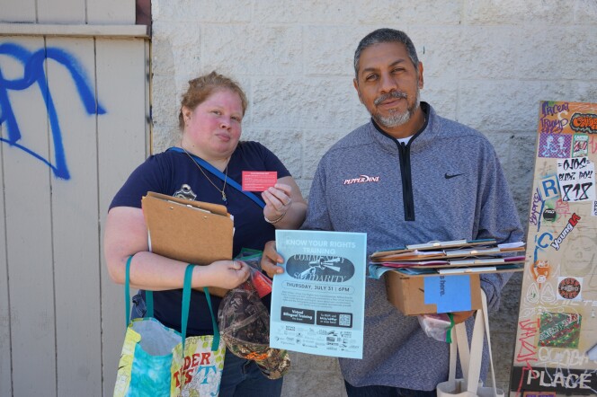 Two people stand together in front of a beige wall with some blue graffiti. They're outside and they stand in the shade. The person to the left is a woman with a light skin tone and light red hair, pulled back. She wears a blue short-sleeved shirt and holds a bag, clipboards and a red piece of paper the size of a business card. To her left, a man with a medium skin-tone and long-sleeved gray zip up holds a blue paper up and holds a stack of clipboards with his other hand.