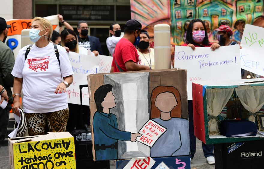 Nearly a dozen people wearing masks stand with homemade signs protesting for tenant protections. In the center a moving box has been painted with a person handing someone with long brown hair a note that reads: "Evicted for failure to pay RENT."