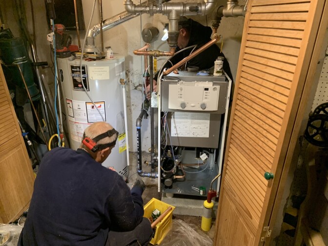 A man with light skin tone wearing headgear is crouched in front of a gas boiler.