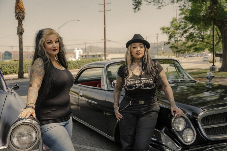 Two Latina women stand in front of their cars. The women are both dressed in black shirts; one wears jeans and the other wears black pants. Both have visible tattoos on their arms and chests, and long black hair that is dyed blond in the front. Their cars are painted black and pristinely clean.