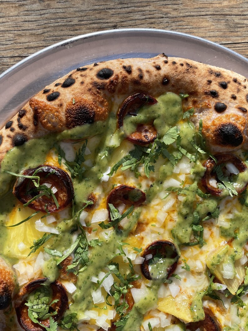A pizza with the outer crust containing a series of black spots from its cooking. The pizza's center contains slices of charred cooked sausage underneath a layer of fresh yellow pineapple covered in a light green salsa and diced white onions.