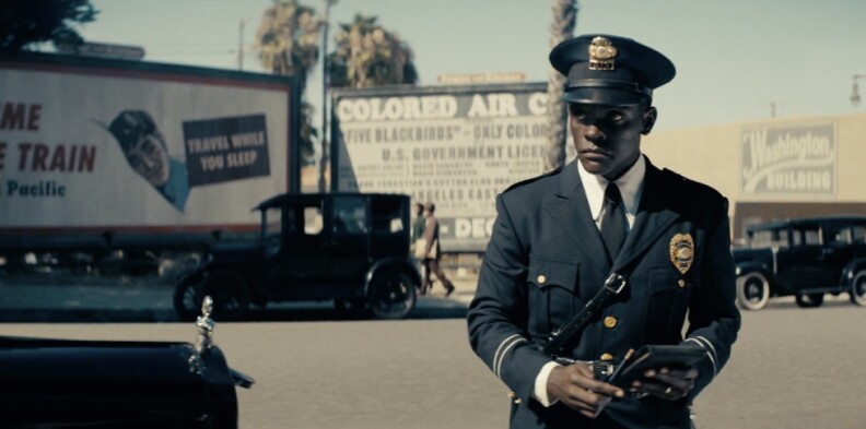 a black man in a police uniform and hat holds his notepad and looks up warily. in the distance behind him are cars and a large billboard that reads "colored air circus"