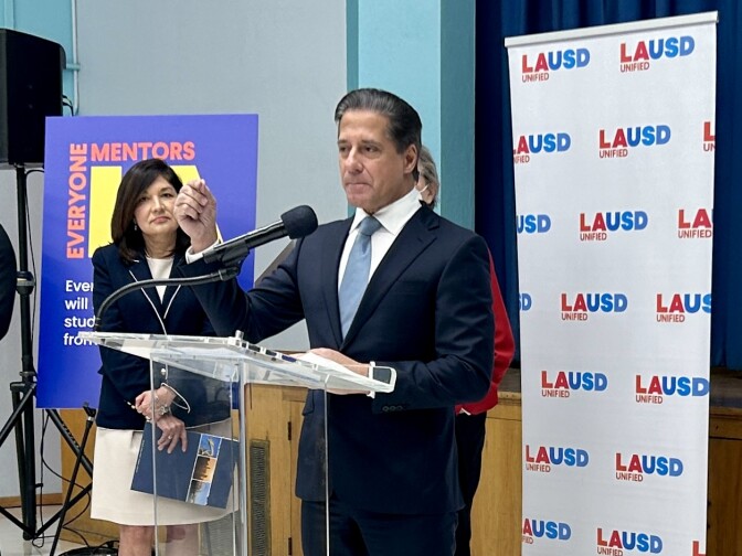 Superintendent Alberto Carvalho stands at a clear lectern with a microphone in front of a portable screen with the letters "LAUSD" printed in red and blue. He wears a navy suit, a light blue tie, and a white collared shirt. His hair is neatly slicked to one side. A woman in a white skirt and a dark blazer stands off to one side with her hands folded in front of her, and she's holding a brochure. Both have a light or olive complexion.