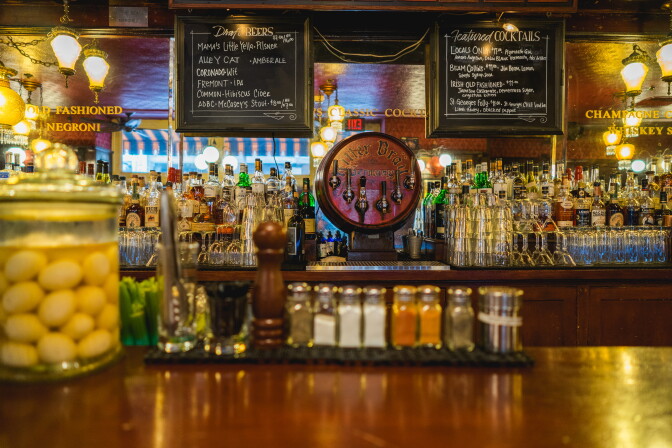 A warmly lit vintage bar interior at Cole’s, showcasing a gleaming back bar lined with glassware, colorful liquor bottles, and a signature “Littler Bros. Fountain” beer tap at center stage; a jar of pickled eggs and a neat row of spice shakers hint at the bar’s old-school charm and cocktail precision.