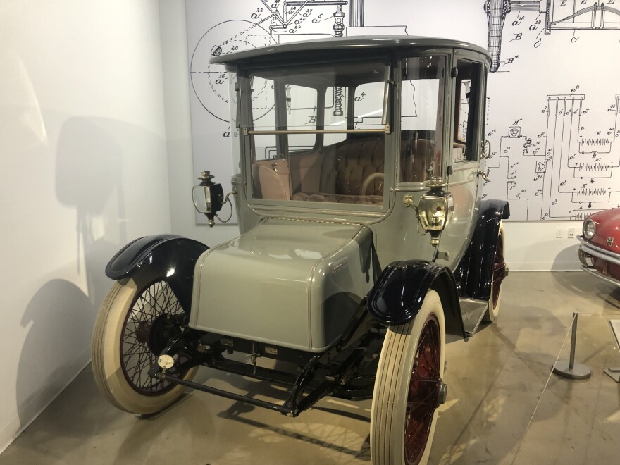 An image of an old car with a light-gray-green finish and white wheels in a museum.