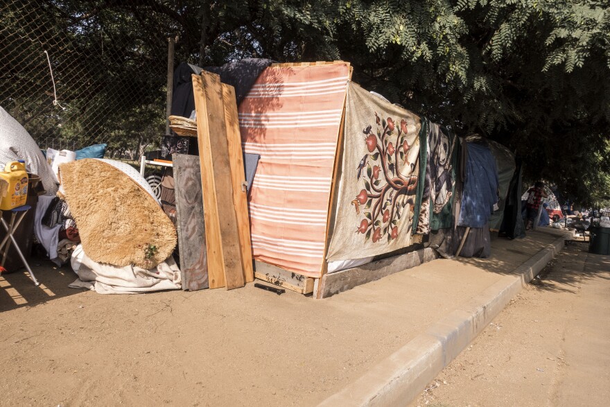 A makeshift lean-to shelter covered in tarps and blankets and surrounded with wood planks and other materials stands on a dusty sidewalk.