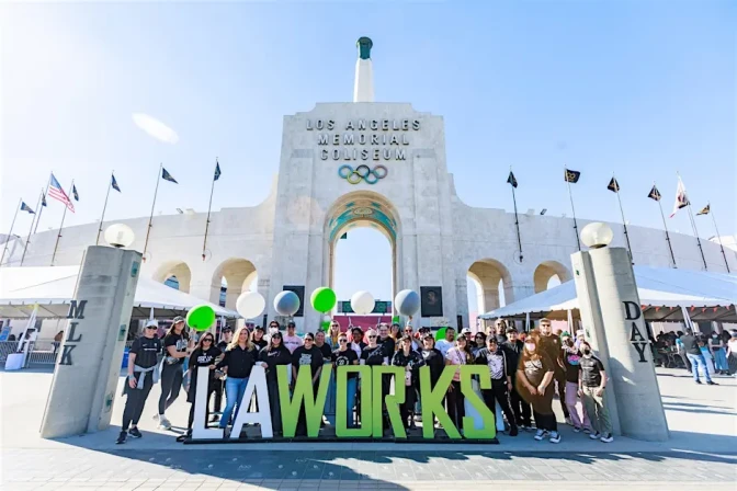 A group of people behind a sign that says"LA WORKS" outside the LA Memorial Coliseum