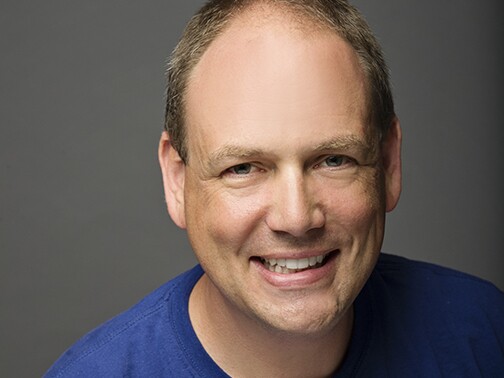 A man in a blue T-shirt with light tan/pinkish skin smiles.