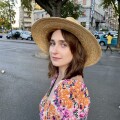 Young woman in a wide brim straw hat, wearing an orange and pink flower dress walks stands in front of a city street.