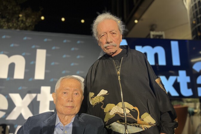 A Japanese American man and Latino man pose together in front signs that read "Am I Next?"