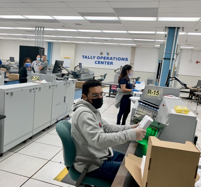 Five people dressed in T-shirts and sweats sit or stand at large scanning machines. 
