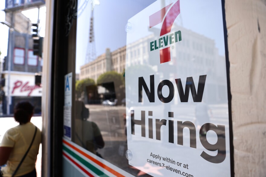 A sign in the window of a 7-Eleven that reads "now hiring. Apply within or at careers.7-eleven.com"