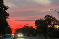 Yesterday's sunset in Los Angeles, California. The sky is red and pink in the background and gets gradually whiter in the foreground. There are trees on either side and cars driving on a road.