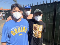 A family of three stands together in front of a black metal gate. From left to right, they include a boy wearing a blue UCLA Bruins shirt, a mother wearing a blue Dodgers baseball hat, and a younger boy wearing a black shirt featuring peace signs. All have medium skin tone and all are wearing masks. 