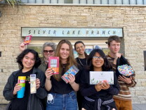 Four teenagers and two librarians stand in front of the Silverlake Branch Library holding art supplies.