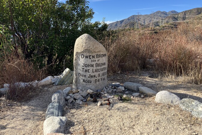 A gray headstone sits on gravelly soil, in front of dry brush and a tree. The headstone reads "Owen Brown, son of John Brown The Liberator, Died Jan. 9, 1889, aged 64 yrs." Smaller stones and pine cones are gathered at the base of the headstone. Mid-sized gray stones mark the ground in front of the headstone into the shape of a grave. The sky is blue and there are hills and telephone lines behind the scene.