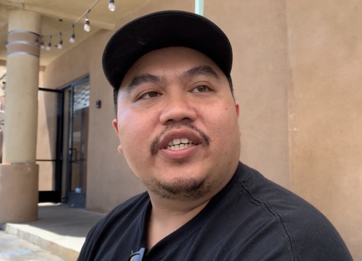 Tony Dang, a founding member of Seniors Fight Back, wears a back T-shirt and ball cap. Behind him a glass door to a strip mall shop is open.