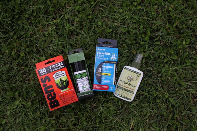 An array of four mosquito repellents on the grass next to each other. From left is the Ben's 30% deet wipes in an orange box. Next is the Junlge Juice spray, which is 99% deet, in its green box. After that is the picaridin lotion from Sawyer in it's blue box. Last is the Herbal Armor spray bottle, which is a light olive green, from All Terrain.