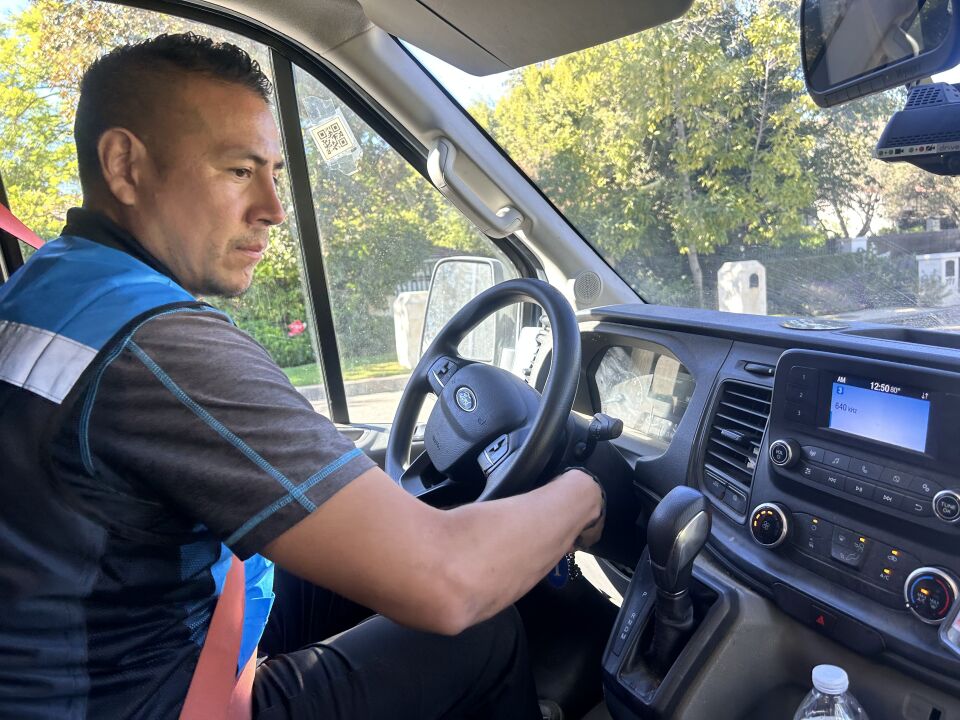 A man wearing a gray t-shirt with a blue work vest overtop is sitting in the drivers seat of a vehicle, his right hand outstretched towards the keys in the ignition. A residential neighborhood can be seen through the windshield.