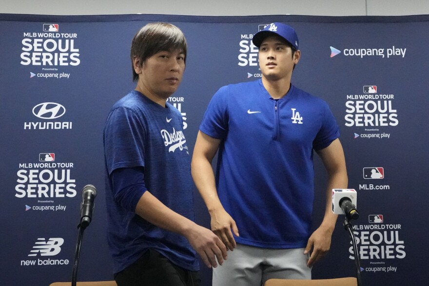 Two Asian men in blue Dodgers shirts stand behind microphones against a blue backdrop.