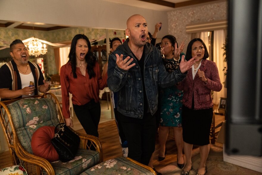 Jo Koy and six other cast members of Easter Sunday stand in a living room, crowded together, making various faces and gestures of anger and disappointment at a television that is just peaking into the frame on the right side of the screen. In the background, living room furniture and a dining room can be seen in the far back.