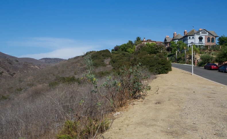 A dirt area sits between a roadway and house and brush on a hill