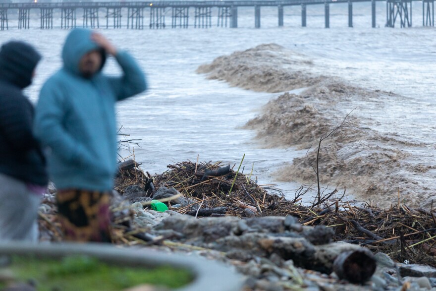 Two men out of focus stand on the shore amidst debris and drift wood, while they look out at the gray and brown ocean water on a stormy day. A pier can be seen in the background at the top of the frame.