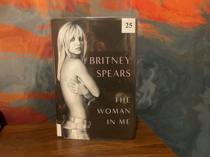 A physical library copy of Britney Spears's memoir "The Woman in Me" against an orange and blue backdrop.