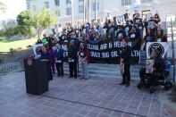 A person speaks at a podium at the steps of L.A. City Hall as a crowd behind her holds banners. One reads"Clean Air & Health Over Big Oil Wealth."