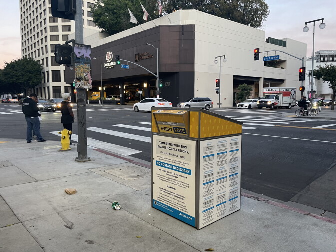 A ballot drop box sits on a street corner. In the background, cars and pedestrians are on the move. 