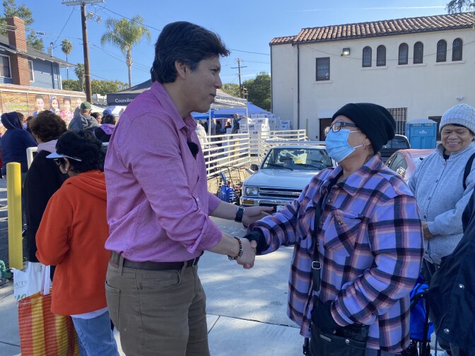 Kevin de Leon, a man with brown skin and dark hair, is standing and shaking the hand of a woman in a mask.