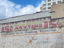 Faded paint on the side of a brick buildin reads "ROXY THEATRE; 15 CENT ANYTIME; GLENDALE'S HOME OWNED THEATER." Some lower faded type reads "Dresses" and "Lingerie." A blue sky is seen in the background, a more modern building to the right.