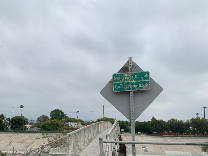 A narrow entrance to the bridge is marked by several metal poles and several bushy grasses. A dinged up sign reads "Sunnynook Bridge" and "Atwater Village 0.5 miles." 