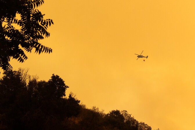 A helicopter is silhouetted against an orange sky due to wildfire.