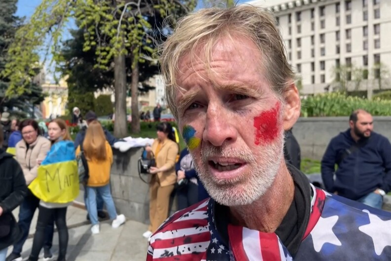 A man with a close-trimmed white beard and red splotches on his cheek appears draped in a U.S.-flag light top.