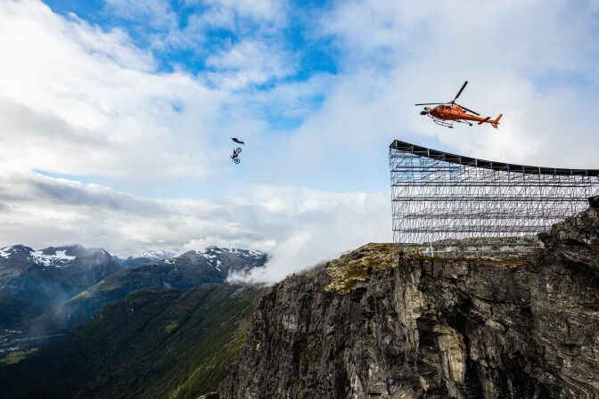 A motorcycle hangs in the air after jumping off a ramp atop a mountain during filming of stunts for the movie Mission: Impossible Dead Reckoning Part One. An orange helicopter follows the motorcycle.