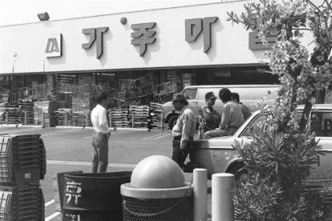 Korean Americans are standing in front of a barricaded Korean supermarket during the LA Riots. 