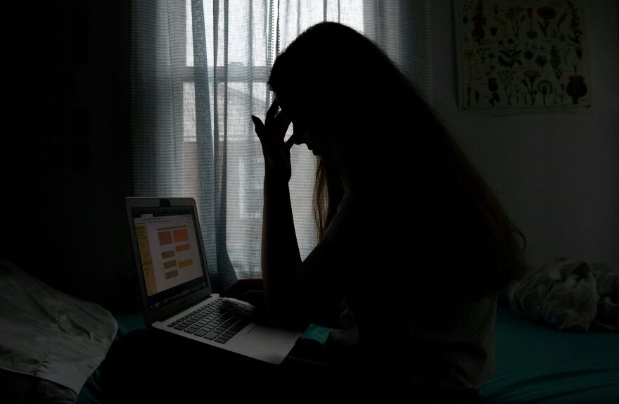A young woman in her teens poses for a picture with a laptop in a darkened room. Her face is dark and obscured by backlighting through a bright window.