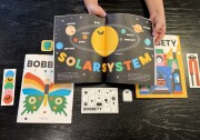 A pair of hands with medium light skin tone hold open a brightly colored magazine depicting the solar system with a bright yellow circle surrounded by smaller circles in different colors. The magazines in the background have a butterfly and ghosts on the cover. 