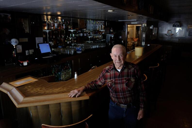A wide shot of a man in a plaid shirt and blue jeans standing inside an empty bar, next to a wooden bar
