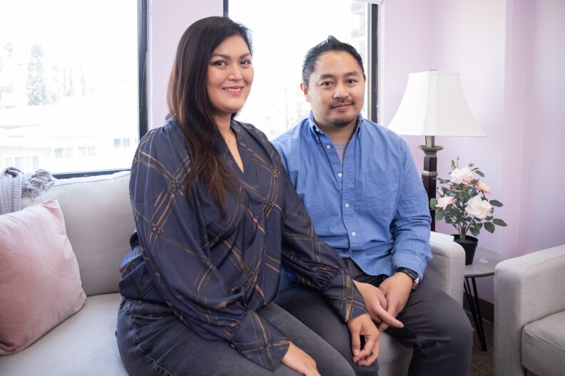 A woman with light skin tone and long dark hair wearing a dark blue blouse sits on a gray couch next to a man with light skin tone dark hair and a blue button up shirt.