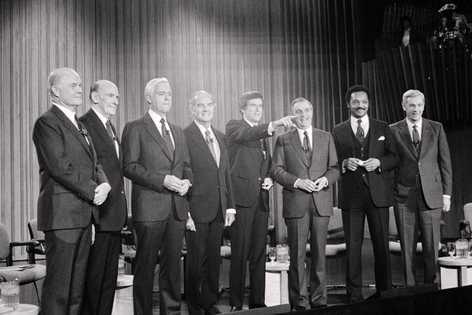 Eight men stand on a stage in suits. Seven men are white, the second man from the right is Black.