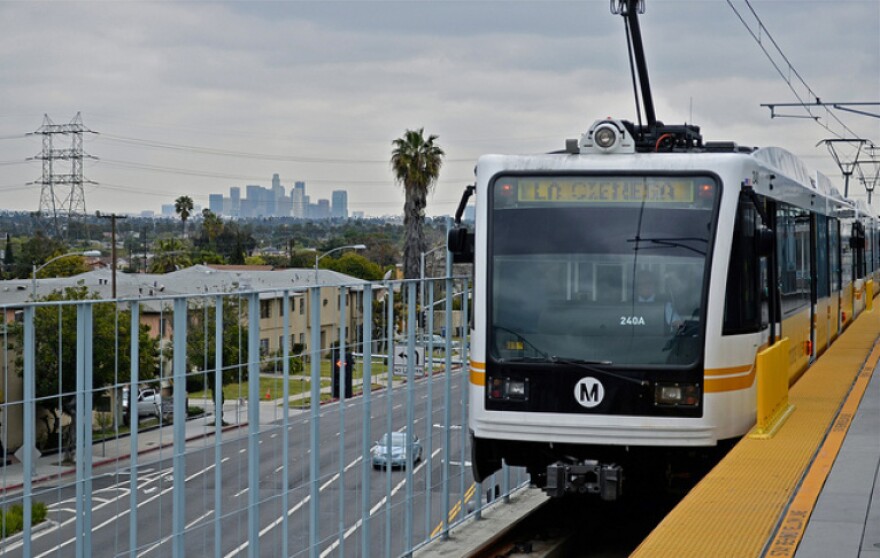 A train arrives at an elevated station as traffic lanes and the downtown Los Angeles skyline are visible in the distance.