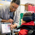 A woman in glasses helps a young boy in a wheel chair with navigating a computer screen.