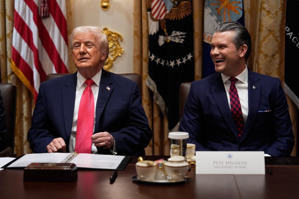 Donald Trump and Pete Hegseth sit next to each other at a table with flags behind them.