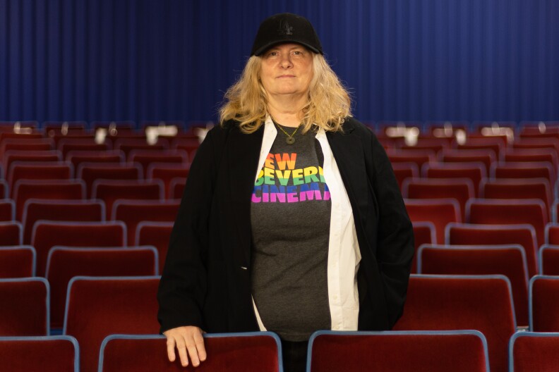 A feminine presenting person wearing a cap, black jacket and t-shirt with the rainbow-colored words "New Beverly" on it stands among the mass of empty seats in a theater, looking at the camera.