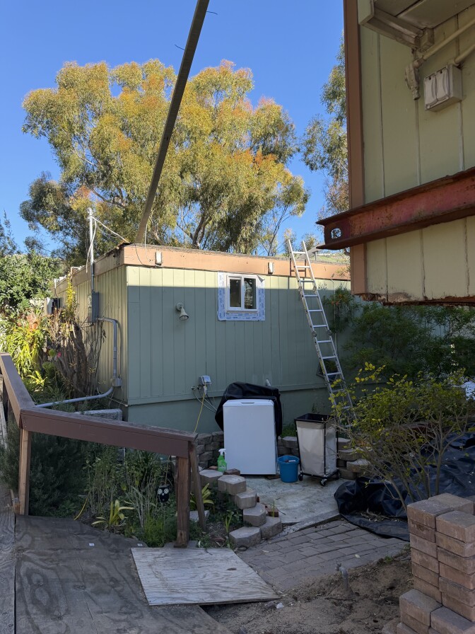 A white washing machine is covered in a yard area of a home. Nearby, a ladder leading to the roof rests against a wall. 