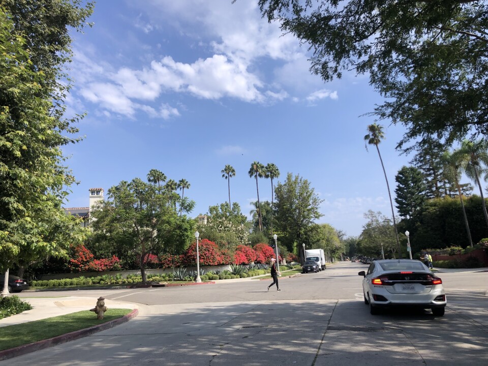A white hybrid vehicle on a shady street, lined with trees and flowers, on a sunny day with blue skies. A woman walks across the street in front of the vehicle. 