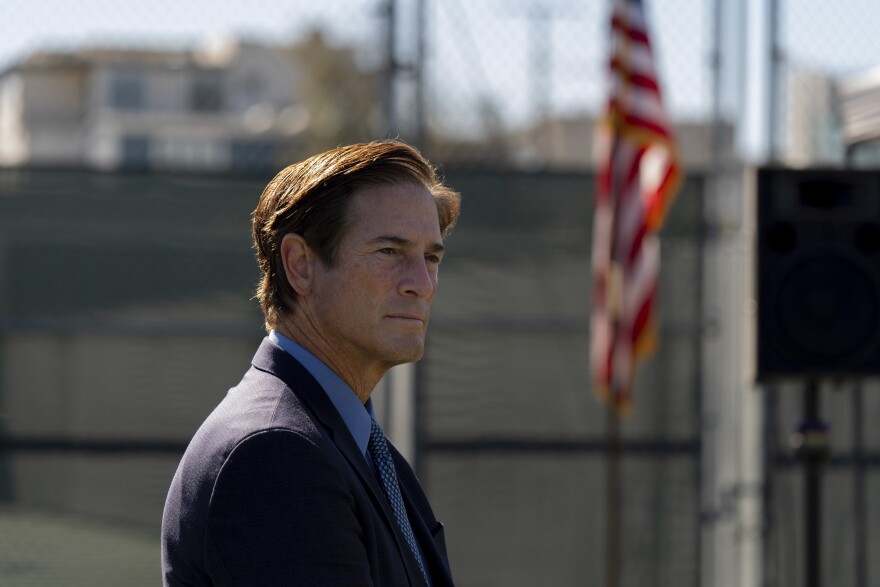 A white man with short hair in a dark blue suit stands outside alone, with a U.S. Flag in the background.