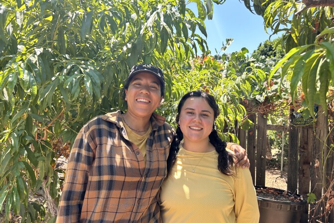 Two women are smiling at the camera with their arms around each other. The woman on the left is wearing an orange long-sleeve flannel and a gray baseball cap, while the woman on the right has her black hair pulled back in a ponytail and is wearing a pale-yellow shirt. They're both standing in an outdoor farm space filled with leaves and trees on a sunny, clear morning.